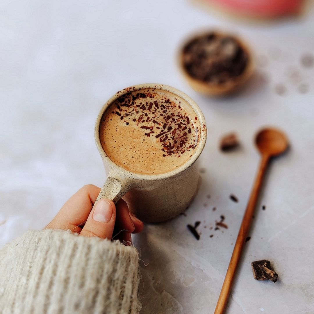 Person holding a mug of Bean and Goose Hot Chocolate with chocolate shavings on a light surface with a wooden spoon.