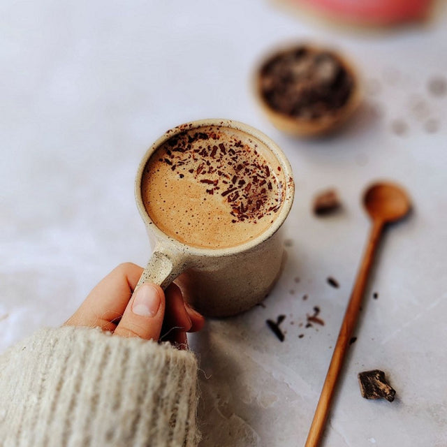 Person holding a mug of Bean and Goose Hot Chocolate with chocolate shavings on a light surface with a wooden spoon.